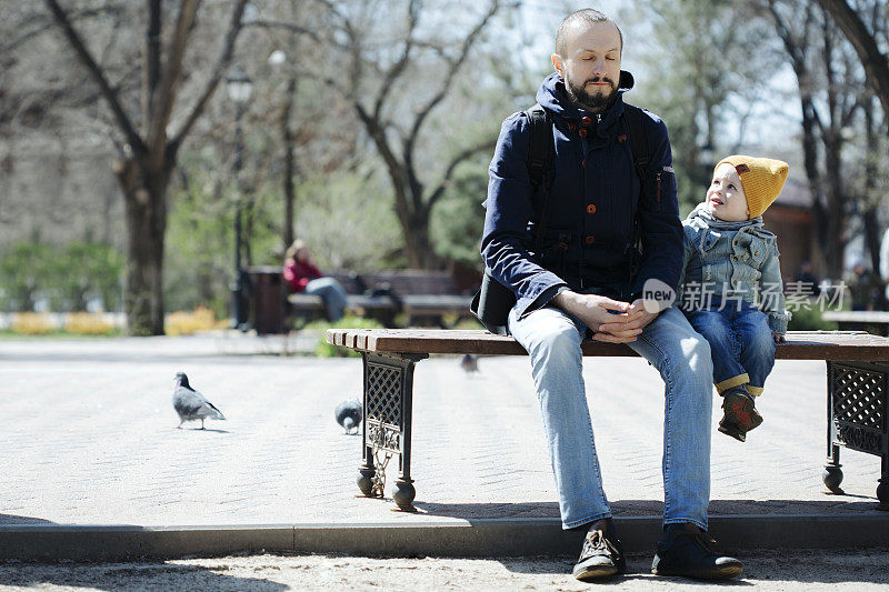 Dad is sitting on a bench in a city park with  son and doing meditation,  his son is surprisingly watching father with interest
